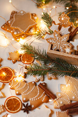 Delicious homemade gingerbread cookies with white icing. Christmas lights, branch of fir tree, dry orange slices. White wooden background, flat lay, top view. Festive holiday atmosphere, family time