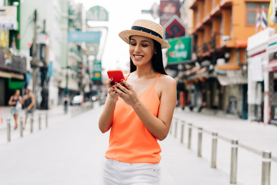 Young brunette woman using a mobile phone laughs in Bangkok, Thailand with attractive smile. Happy and excited Hispanic girl traveler wear summer clothes with hat in Asia. With Copy space