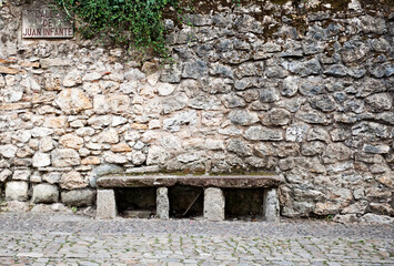 Sitzbank vor einer Natursteinmauer in einer gepflasterten Gasse (Calle de Juan Infante) in Santillana del Mar, Cantabria; Kantabrien; Spanien;