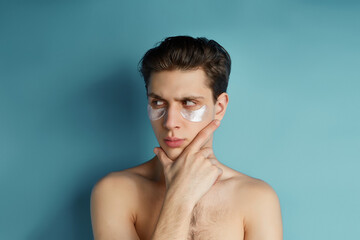 Fototapeta na wymiar Busy men prefer minimalist, easy and quick facial treatments. A young brown-haired man with white gel patches under his eyes is scowling and looking away.