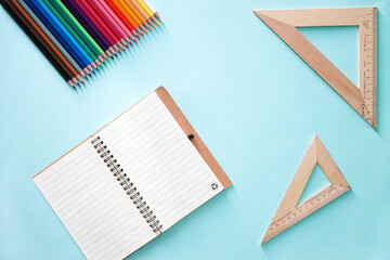 Horizontal flat lay composition with Clear sheet note book, two wooden rulers triangles and colour pencils on a light blue background. Back to school, college, education concept.. Art supplies idea.