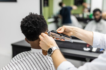 Barber trim hair with clipper on young unshaven black man in barbershop studio. Professional hairdresser cut hair with electric shearer machine on African guy. Male beauty treatment process