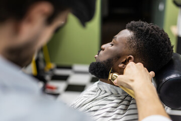 Barber trim hair with clipper on young unshaven black man in barbershop studio. Professional...
