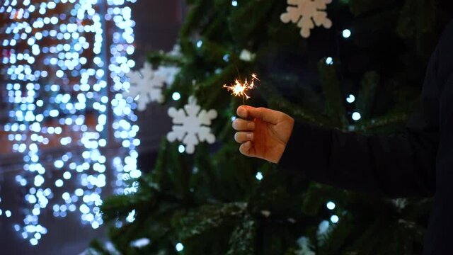 Sparkler burns out and goes out in one male hand at night against the background of lights and a Christmas tree, close-up