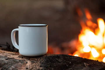 Fototapeta na wymiar camping in the woods. White camping cup with tea on a background of fire. summer camping. relaxation in nature