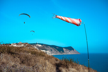 A red and white striped windsock on the seashore amid two flying paragliders. Two paragliders fly...