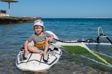 Obraz premium Boy sits on a windsurfing board on the waves of the sea against the sea horizon. sail of windsurfing board lies in water