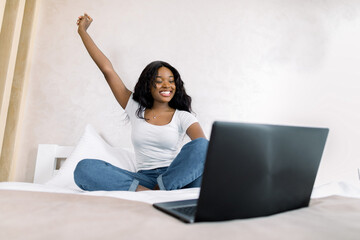 Portrait of smiling young African woman using laptop in bed at home, having video call with her friend, realxing with arm raised up. Good mood on quarantine, lifestyle concept