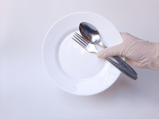 A hand with white latex glove holds a metal spoon, a fork, and an empty plate. Isolated on a white background.