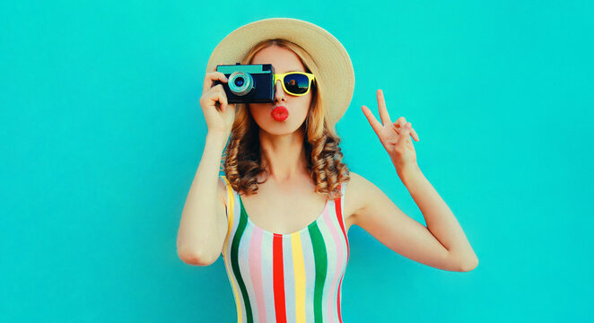 Portrait of young woman photographer with vintage film camera blowing red lips sending sweet air kiss wearing a summer straw hat over blue background