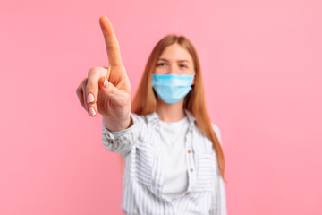 young woman in a medical protective mask on her face, touching an imaginary touch screen with her finger, on an isolated pink background