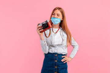 smiling young woman in a medical protective mask on her face, standing with a camera isolated on a pink background. Travel, tourism, coronavirus,