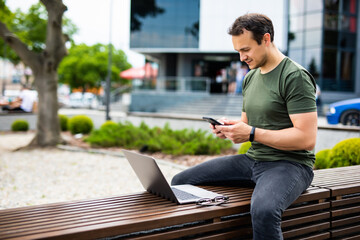 Attractive confident young man sitting on a bench outdoors at the city street, working on laptop computer, using mobile phone