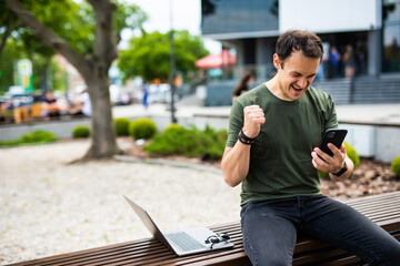 Obraz premium Happy young man celebrate win look at phone sitting on a bench in the park