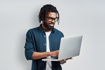 Happy young African man in eyewear using laptop while standing against grey background