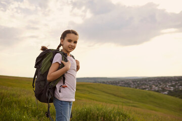 Adorable little girl with backpack standing on mountain top, copy space. Cute kid with rucksack hiking in countryside
