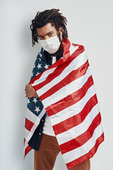 Handsome young African man covered with American flag wearing medical face mask and looking at camera while standing against grey background