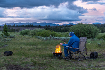 A Camper Man sitting on a rocking chair warming his hands by the fire, views of distance mountains and dramatic clouds over fields with grass, shrub and forest, Yellowstone National Park, Montana