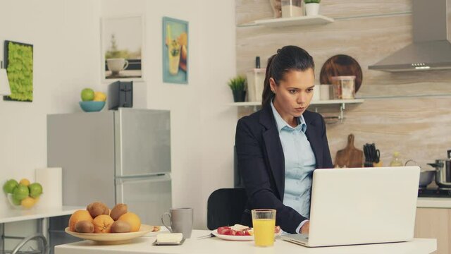 Business woman eating toasted bread with butter while working on laptop during breakfast. Concentrated business woman in the morning multitasking in the kitchen before going to the office, stressful