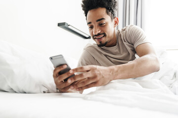 Photo of smiling african american man using mobile phone in white bed