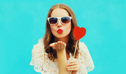 Portrait beautiful young woman with red heart shaped lollipop blowing lips sending sweet air kiss on blue background