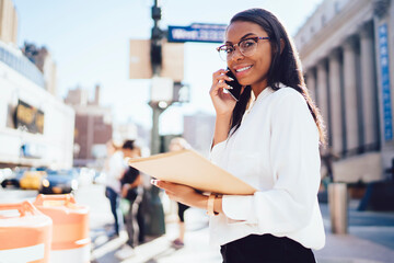 Portrait of cheerful dark skinned female employee holding folder in hands while calling with friends on smartphone device standing in urban setting.Successful lawyer in white shirt talking on cellular