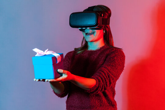 Happy Girl In VR Glasses Holding Wrapped Gift Box And Smiling Joyfully, Giving Christmas Present To Virtual Friend, Celebrating Holidays Birthday In Augmented Reality. Colorful Neon Light Studio Shot