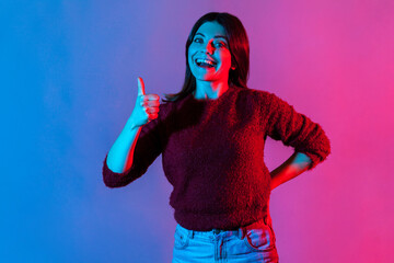 Well done, good job! Neon light portrait of joyful brunette woman showing thumbs up, like gesture to camera and smiling broadly, expressing approval, excellent feedback. indoor studio shot isolated