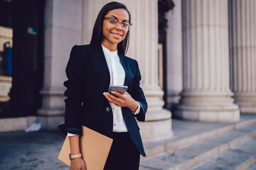Portrait of successful African American businesswoman dressed in stylish suit holding in hand folder and mobile phone while standing outdoors near financial office, young woman lawyer using smartphone - Powered by Adobe