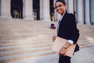 Half length portrait of positive woman executive dressed in elegant suit standing in city outdoors. Cheerful African American lawyer holding folder with documents standing against courthouse building