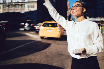 Happy successful businesswoman with dark skin in eyewear catch yellow taxi on street of modern city.Positive afro american female with digital smartphone waving hand standing in traffic movement