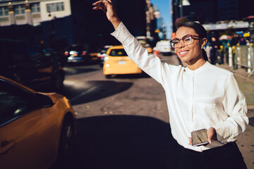 Happy smiling dark skinned woman waving with hand for catching yellow cab and getting to destination in time, cheerful female in eyewear for vision correction hailing taxi in financial district