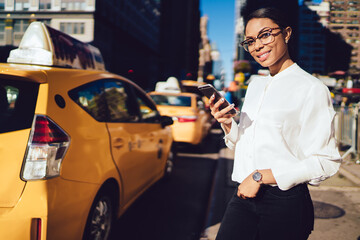 Half length portrait of cheerful young dark skinned woman in casual apparel calling taxi via...