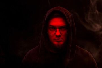 A warlock with a red face in a hood and glasses on a black background in smoke.