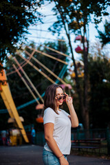 a girl with dark glasses takes off and puts them on the background of the Ferris wheel in the park. girl in a white T-shirt, and the Ferris wheel of different colors