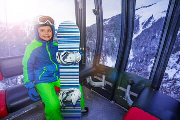 Little happy snowboarding boy with snowboard sit in ski lift cabin lifting on the mountain