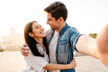 Image of smiling multicultural couple hugging and taking selfie photo