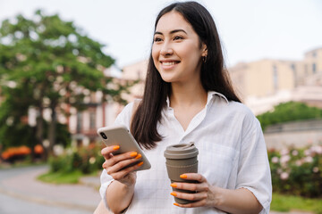 Photo of happy asian woman using cellphone and drinking coffee takeaway