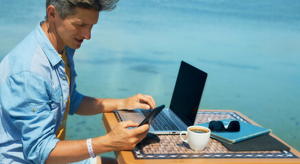 man freelancer working on the beach by the sea, using smartphone and laptop computer. Working and traveling. Freelance concept