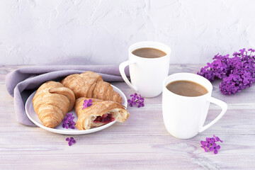 Two beautiful porcelain cups of coffee with milk with croissants decorated with lilac flowers on white wooden table. Perfect breakfast concept. Copy space.