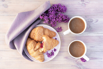 Two beautiful porcelain cups of coffee with milk with croissants decorated with lilac flowers on white wooden table. Perfect breakfast concept. Flat lay style.