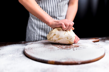 Woman kneading dough on wooden board sprinkled with flour with a black background. Concept of making dough for pizza. Close-up lock of a piece of dough being kneaded by a chef.