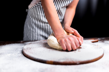 Woman kneading dough on wooden board sprinkled with flour with a black background. Concept of making dough for pizza. Close-up lock of a piece of dough being kneaded by a chef.