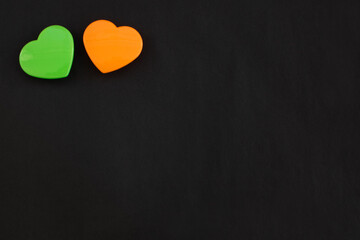 Fototapeta premium Two green and orange color hearts lying close together isolated at the top left corner of black paper background. Flat lay with empty space for text.