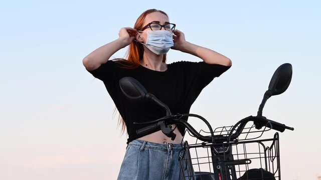 caucasian girl stand near red electro bicycle at background of blue sky puts on medical mask, looks in the mirror. lifestyle, coronavirus, health protection, quarantine, safety and pandemic concept