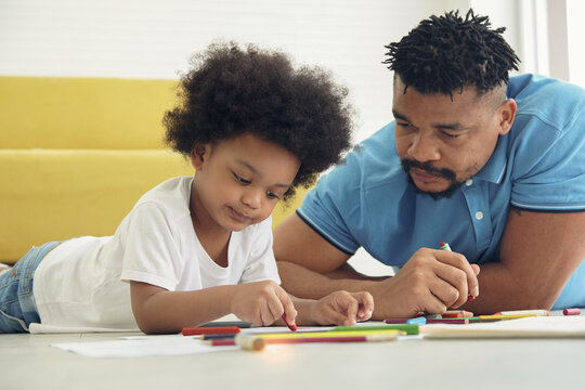 Black African American family dad son together at home. Young father lying on the floor and watching kid child boy drawing coloring pictures
