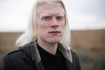 Portrait of a young, beautiful albino man with blue eyes and shoulder hair in a black quilted hooded coat, looking at the camera with a shrewd look against the background of the field. - obrazy, fototapety, plakaty