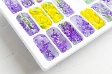 Ice cubes with yellow and violet flowers in a tray close-up. Bright hues.