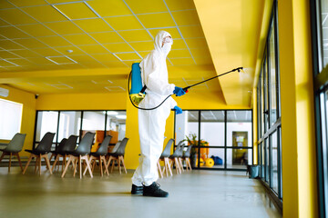 Contractor disinfecting office for COVID-19 coronavirus. A disinfector man in a protective suit and mask sprays disinfectants in office. Prevention of spreading pneumonia virus with surfaces.