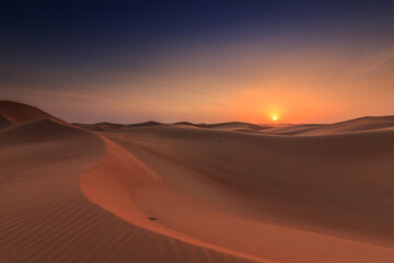 Sunset in the wahiba sands desert of the Sultanate of Oman. Untouched and endless dunes in the evening glow orange from the evening sun. Blue sky with sun and few clouds on the horizon